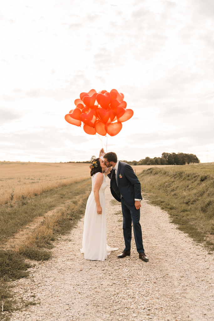 Brautppar küsst sich auf einem Schotterweg und hält Herz rote Luftballons in der Hand.
