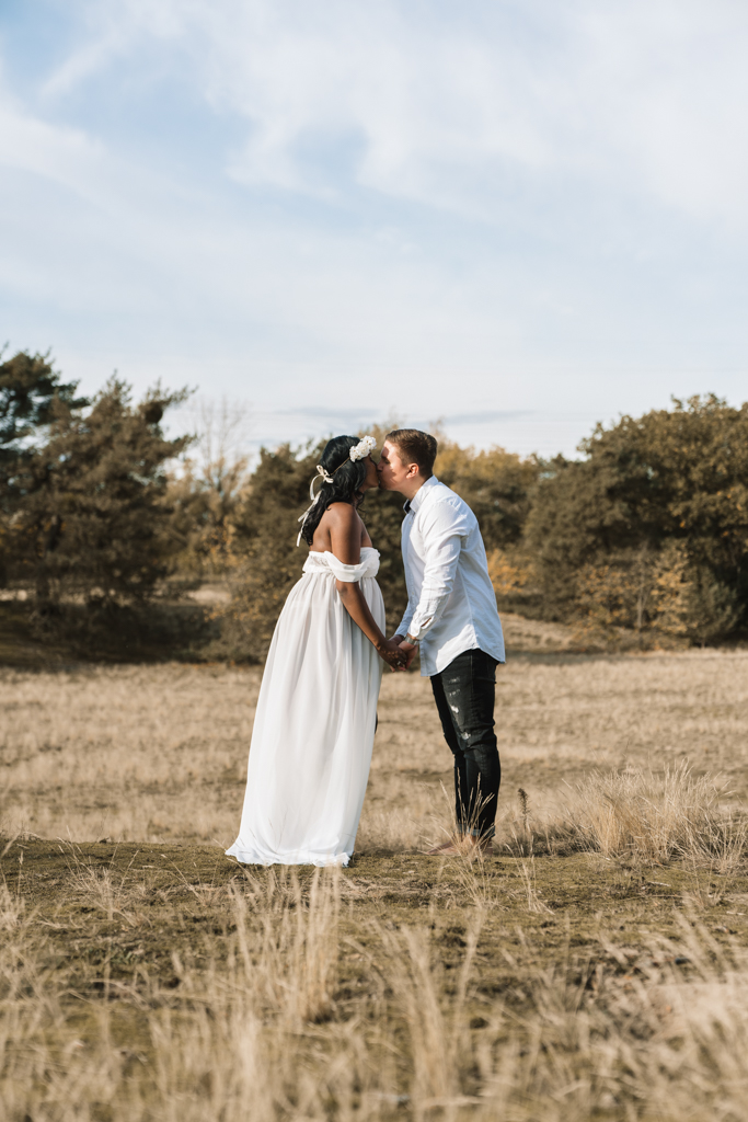 Pärchen küsst sich auf einer Wiese im weißen Kleid und blauem Hemd.