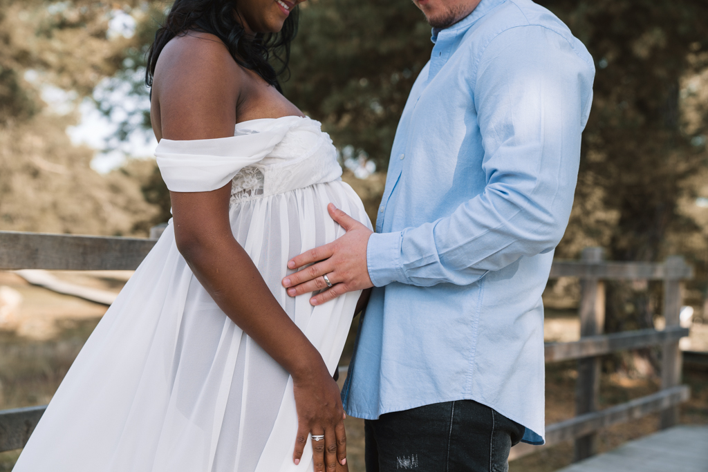 Papa hält den Babybauch im weißen Kleid sanft fest.