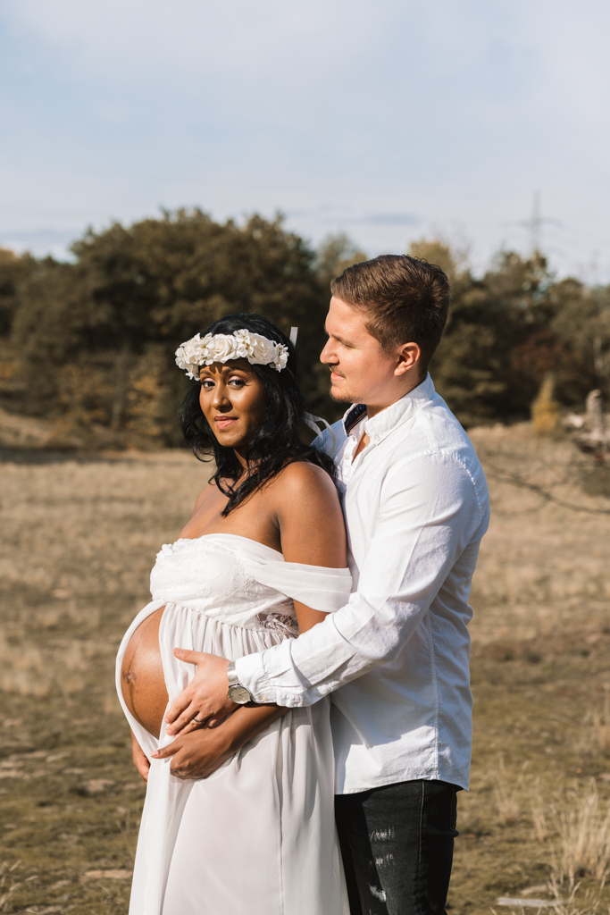 Papa hält von hinten den Babybauch seiner Frau im weißen Kleid.