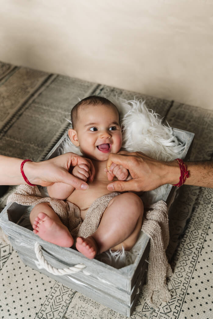 Baby liegt in einer grauen Fotokiste und hält lachend die Hände der Eltern.