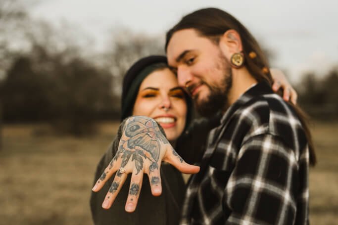 Frau mit Tattoos hält ihre Hand mit dem Verlobungsring in die Kamera.
