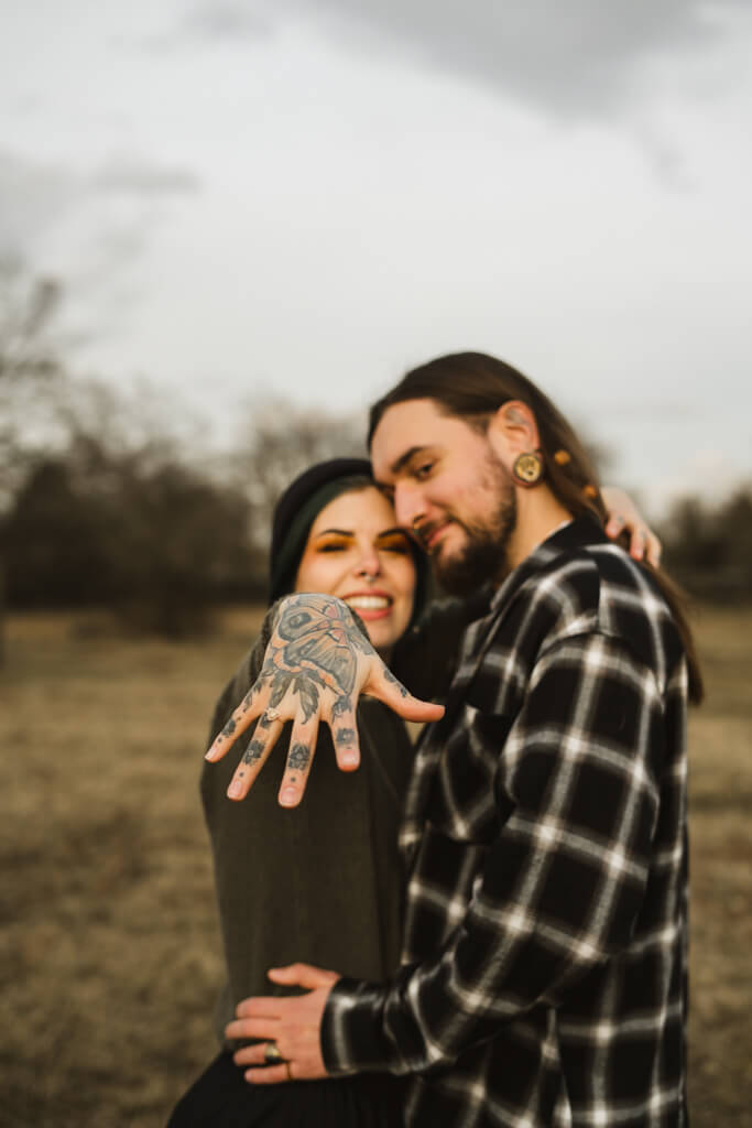 Frau mit Tattoos hält ihre Hand mit dem Verlobungsring in die Kamera.