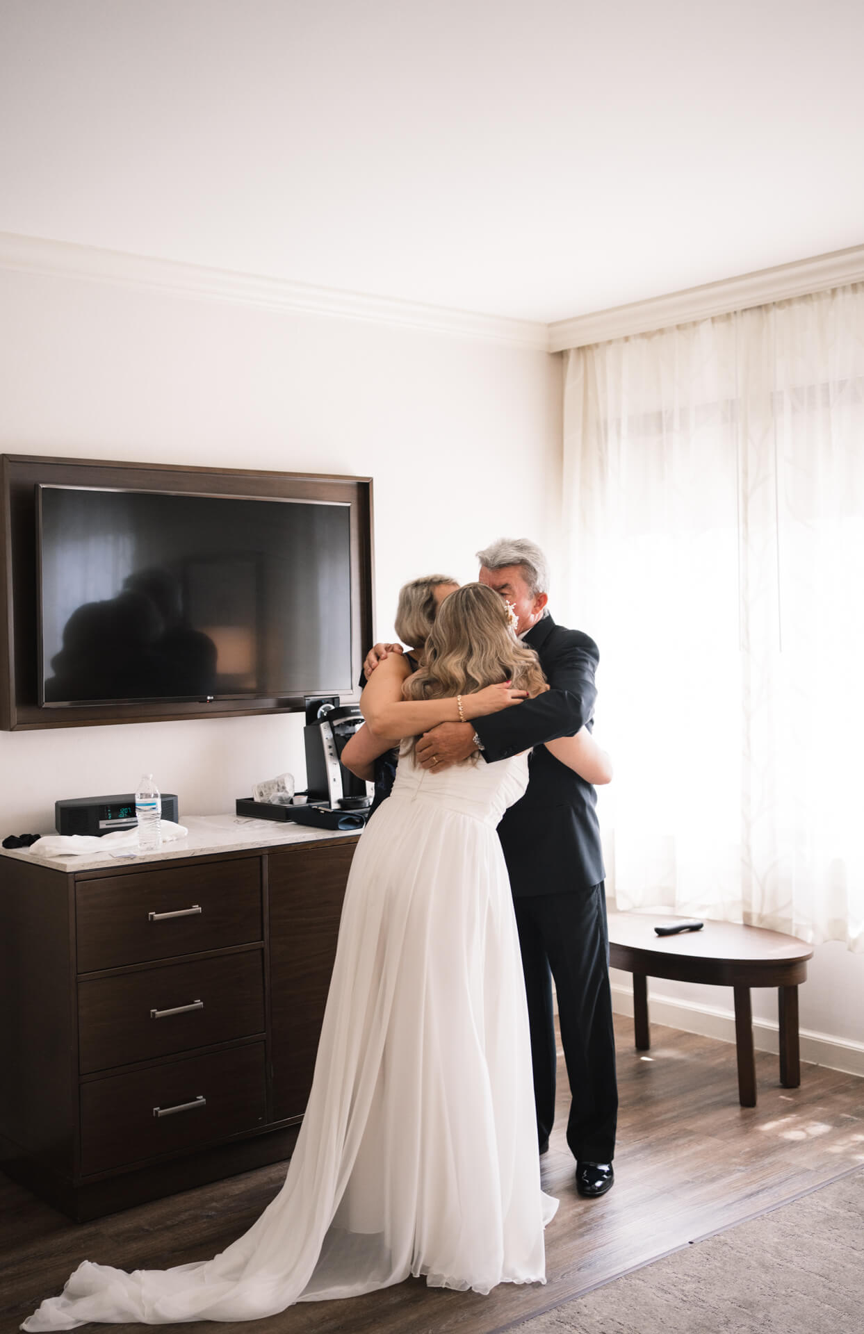 Braut umarmt ihre Eltern in einem Hotelzimmer.