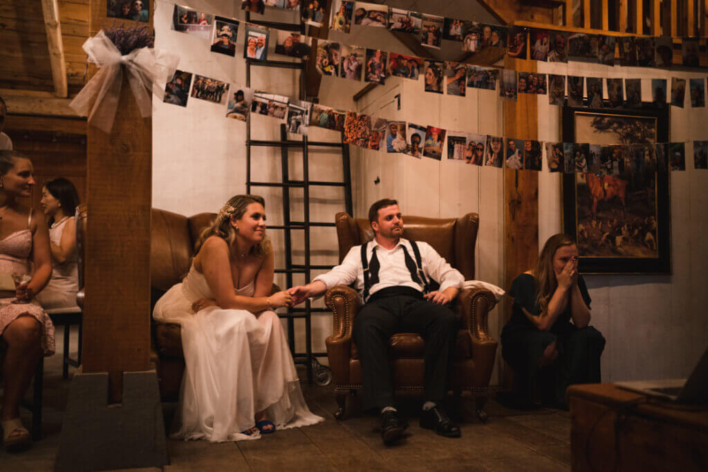 Brautpaar sitzen in Ledersesseln, halten Händchen und schauen gespannt einer Diashow zu.